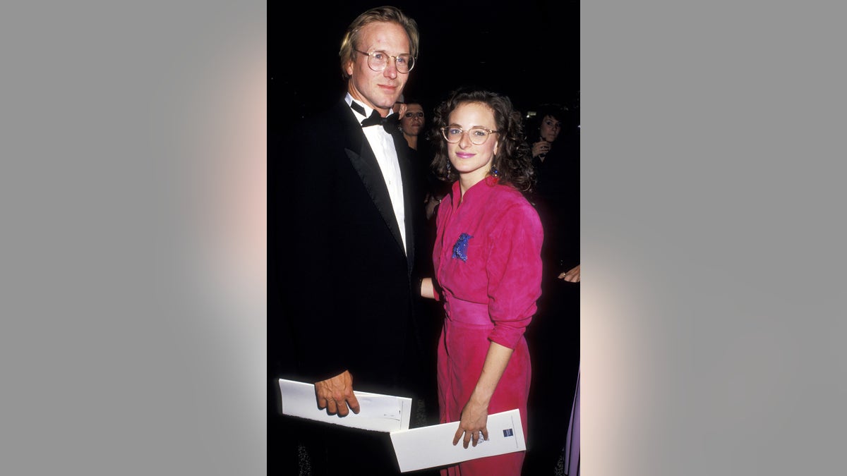 William Hurt in a suit and bowtie standing next to Marlee Maitlin in a hot pink dress and glasses.
