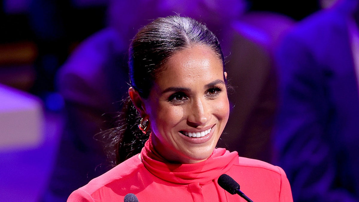 A close-up of Meghan Markle smiling wearing a high collar red suit in front of a podium