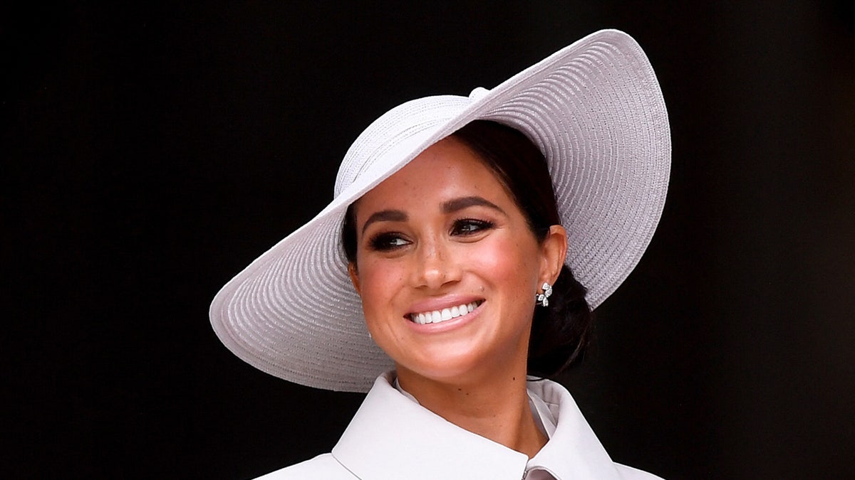 A close-up of Meghan Markle wearing a white coat dress and smiling with a matching hat.