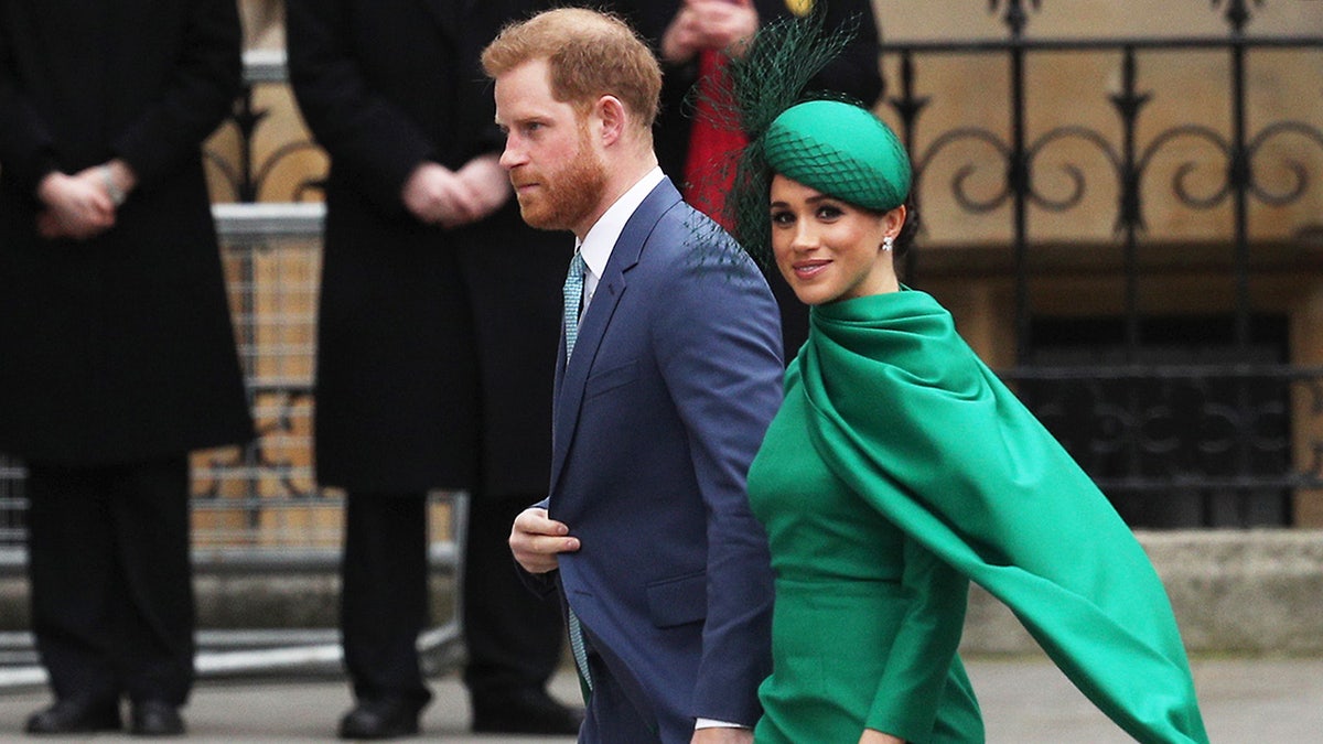 Prince Harry walking away in a blue suit with Meghan Markle in a green suit and matching hat and cap smiling.