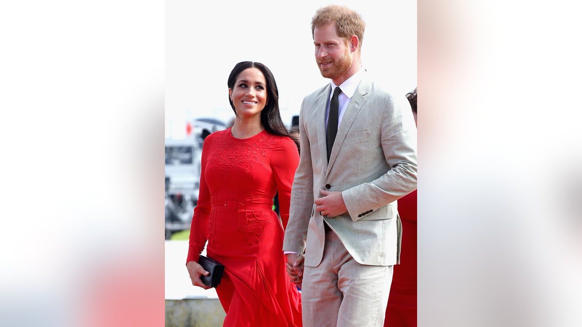 Megan Markle wears a red dress in Prince Harry wearing a gray suit and a dark tie.