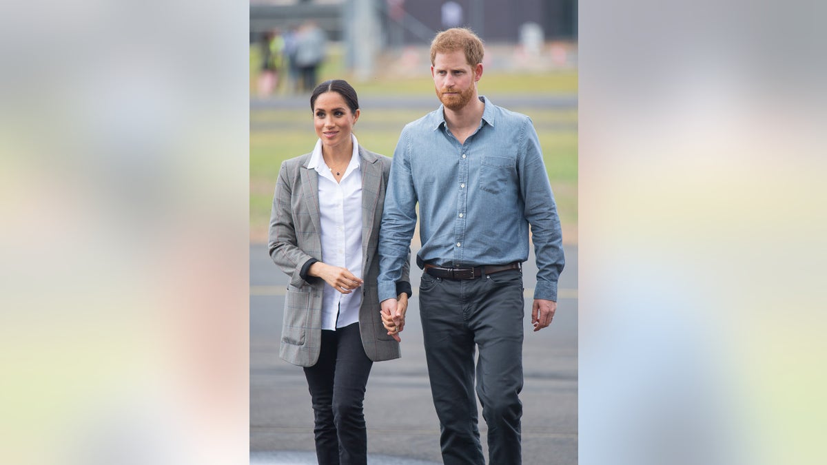 Megan Markle wears a white blouse and a gray jacket walking next to Prince Harry wearing a blue shirt and dark blue pants.