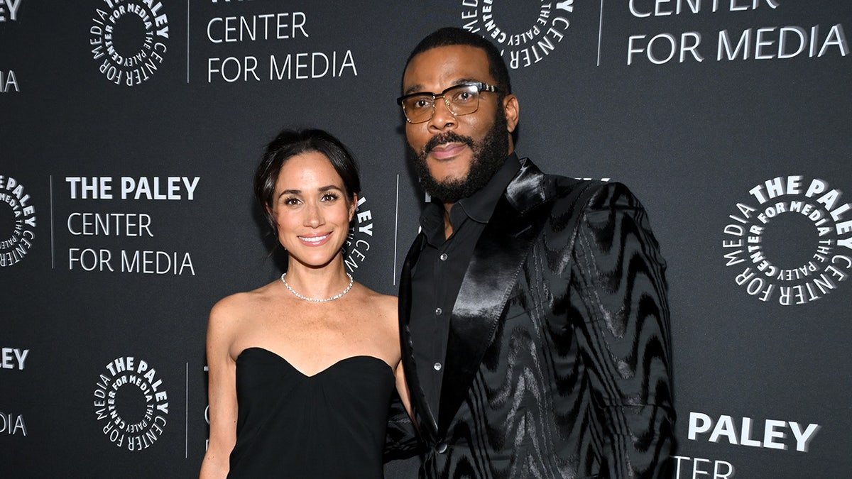Megan Markle wears a black dress without bras standing next to Tyler Perry wearing all black.