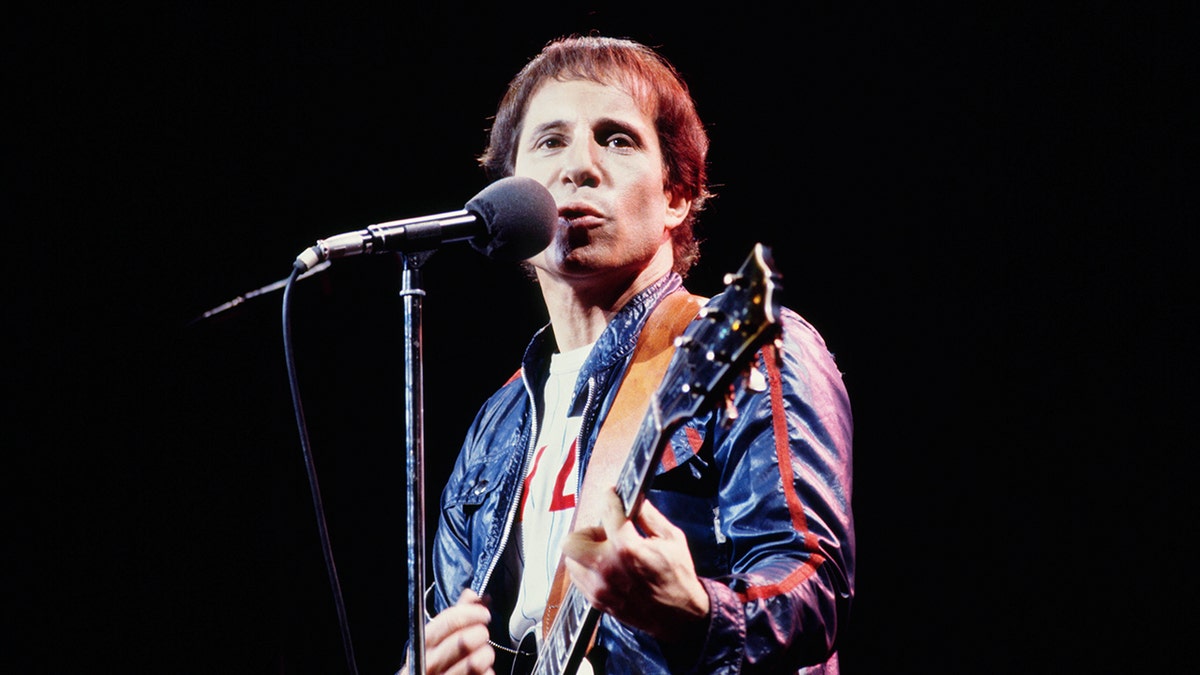 Singer Paul Simon wears a blue jacket and holds a guitar on stage.