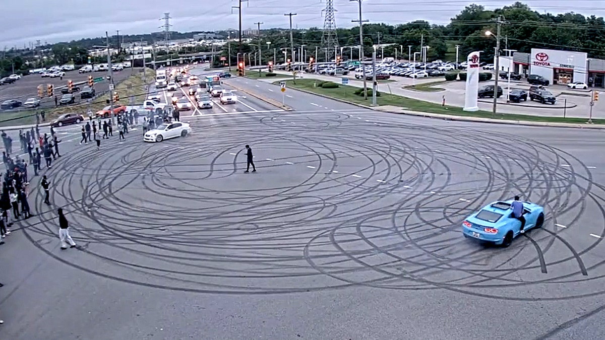 a large crowd surrounds intersection during street takover as cars make multiple donut marks on street