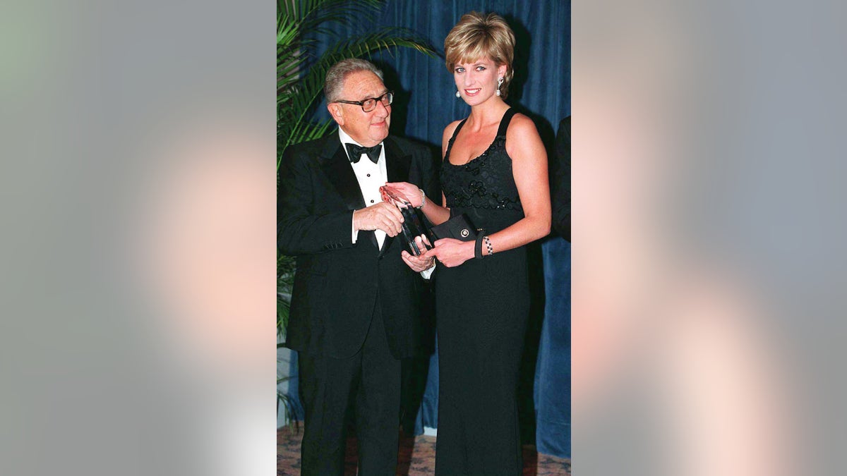 Princess Diana wearing a sparkling black strappy dress posing next to an older man in glasses wearing a suit and bow tie.