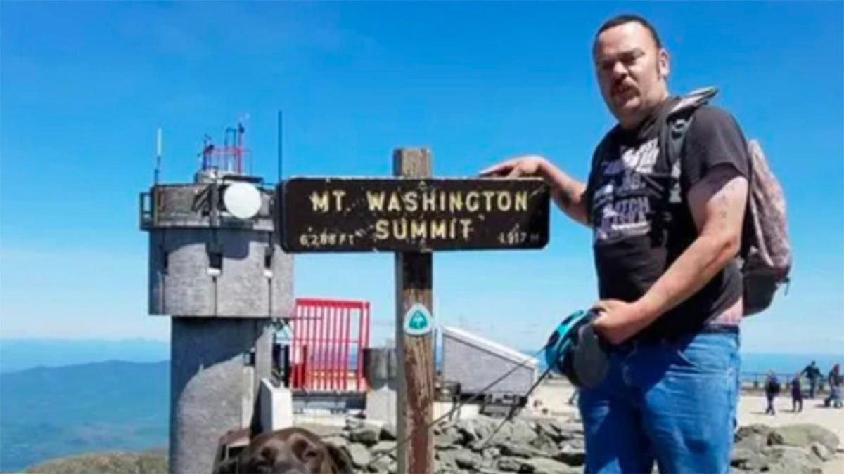 russell hardy standing at the summit of Mt. Washington in blue jeans and a black t-shirt