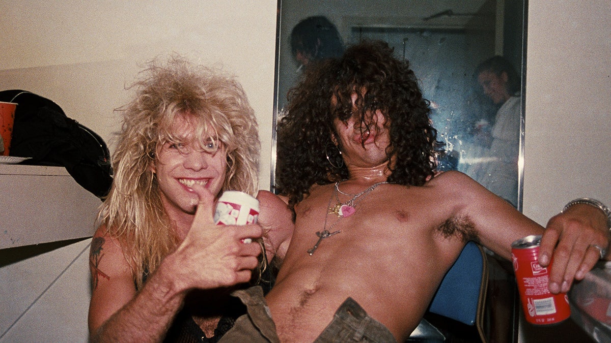 Steven Adler giving a thumbs up leaning against a shirtless Slash.