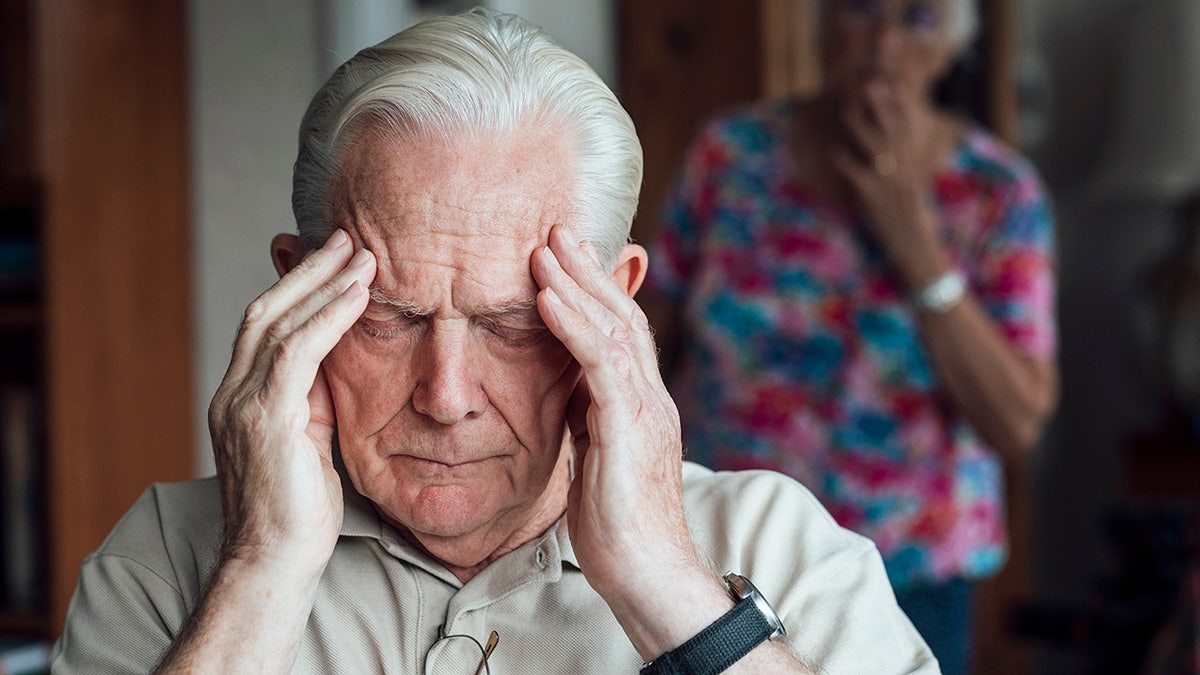 A senior person who has a chronic disease/Alzheimer's disease sitting in his living room in his home in the northeast of England. It keeps his forehead with a negative expression, feeling anxiety/pain. His wife does not focus behind him, looking at him.