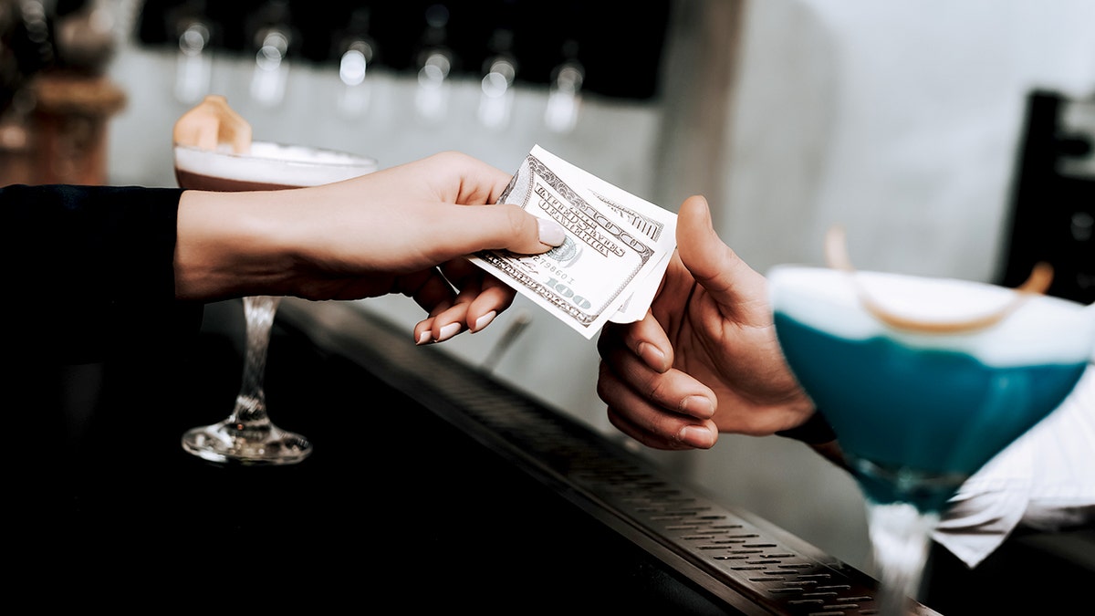 A woman's hand holds a $100 bill for a bartender between two martini glasses.
