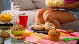 Woman eating on couch surrounded by colorful junk food like cupcakes and chips.
