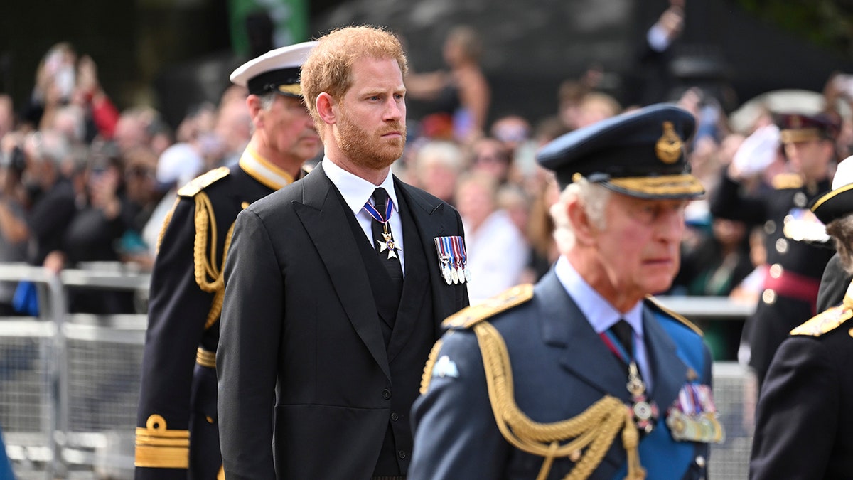 Prince Harry in a black suit walking behind his father who is wearing a blue uniform.