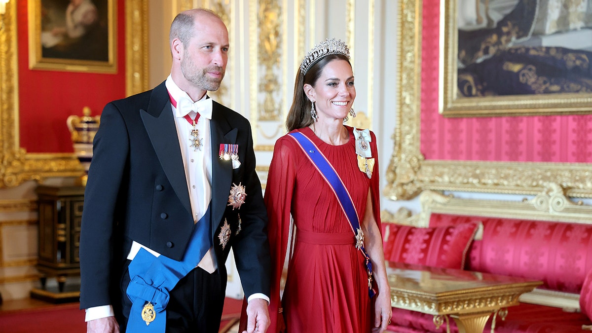 Prince William in a black suit with medals walking next to Kate Middleton wearing a red gown and a blue sash with a tiara.