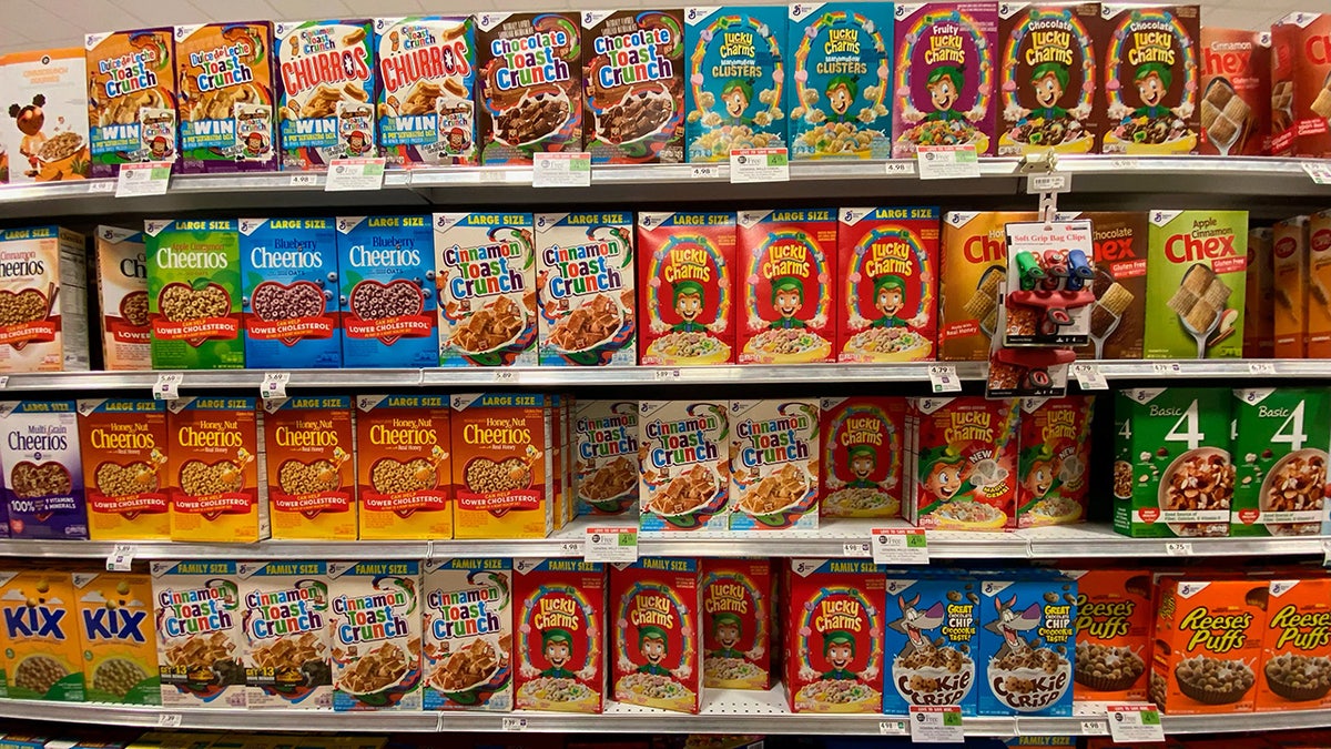 Rows of cereal boxes lined up on shelvers in grocery store. Cinnamon Toast Crunch, Lucky Charms and Cheerios among selection.