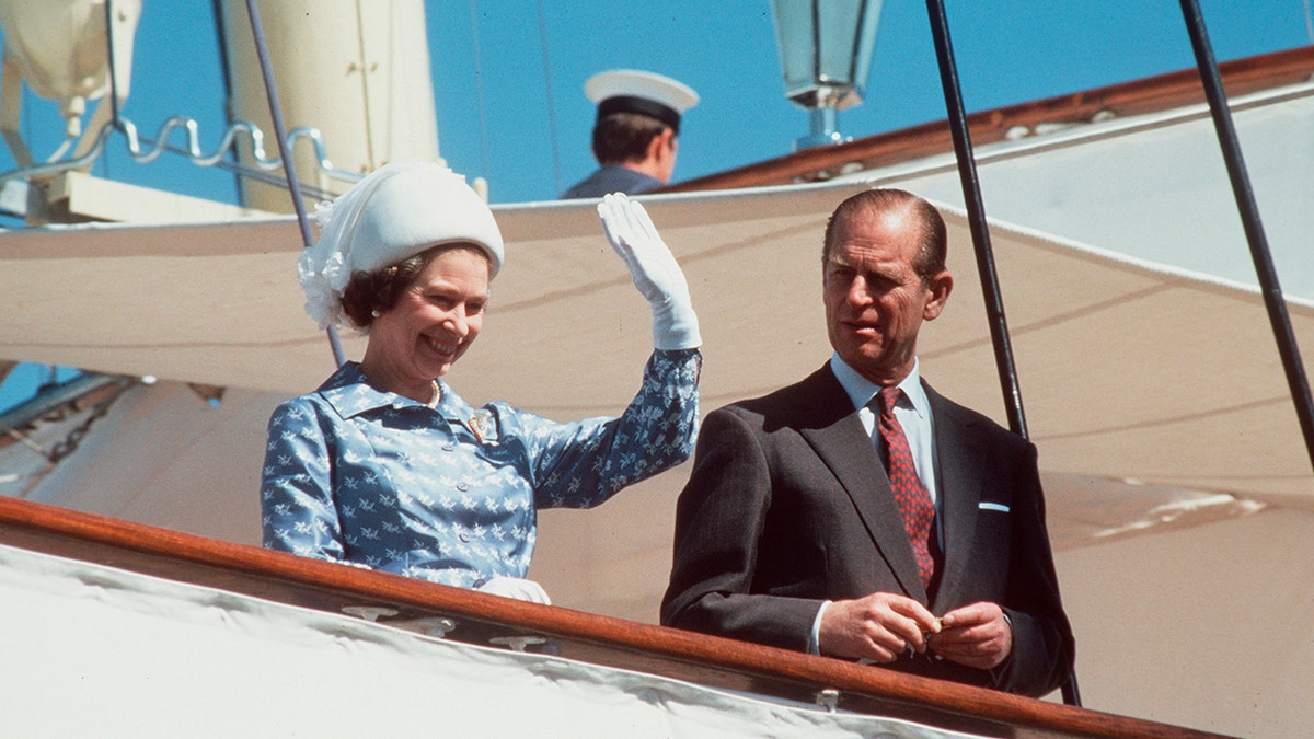 Queen Elizabeth waving in a light blue coat dress as Prince Philip looks on.