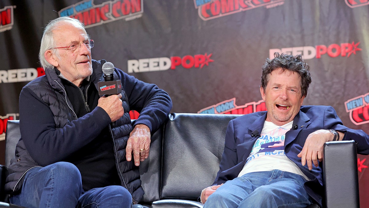 Christopher Lloyd and Michael J. Fox sitting on a couch next to each other and smiling.