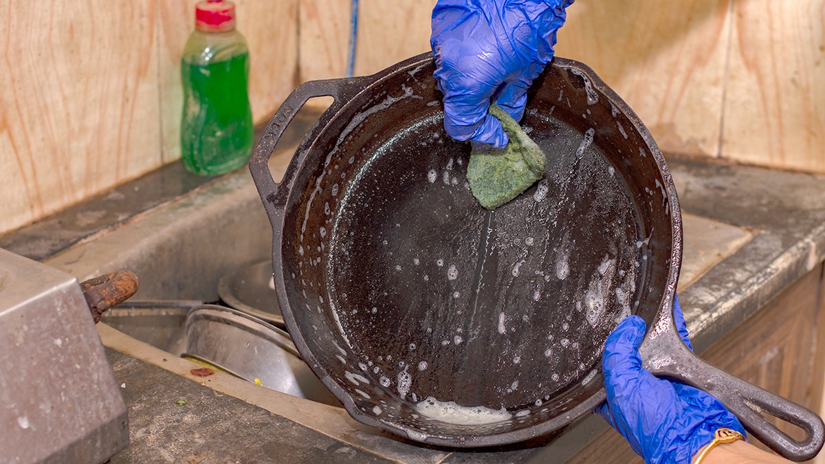 Hands covered with gloves use a scrubber to clean a cast-iron pan.