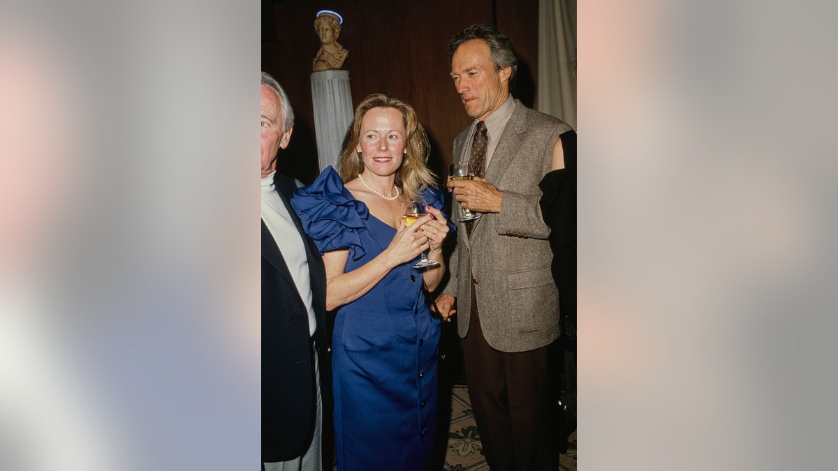 Clint Eastwood, wearing a grey tweed blazer over a beige shirt with a brown polka dot tie, and Jacelyn Reeves, who wears a blue ruffle sleeve dress