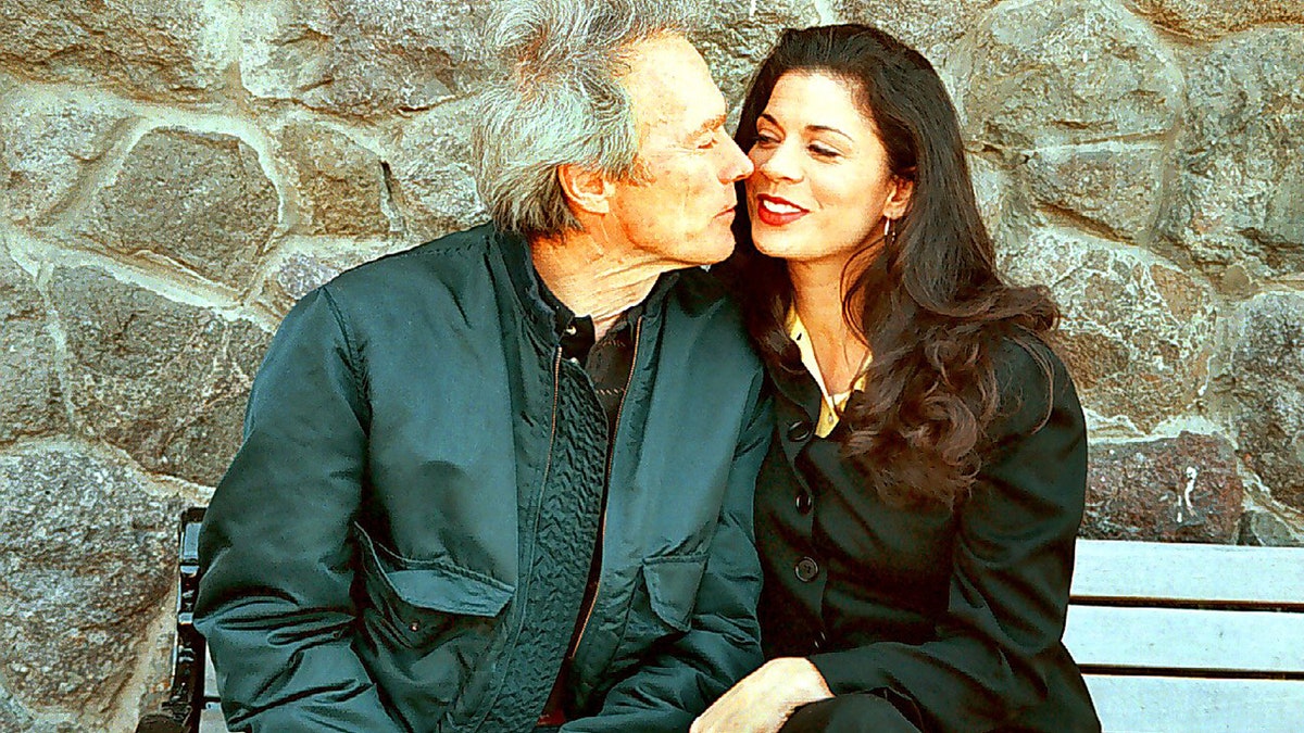 Clint Eastwood wearing a hunter green jacket and shirt kissing Dina Eastwood wearing all black.