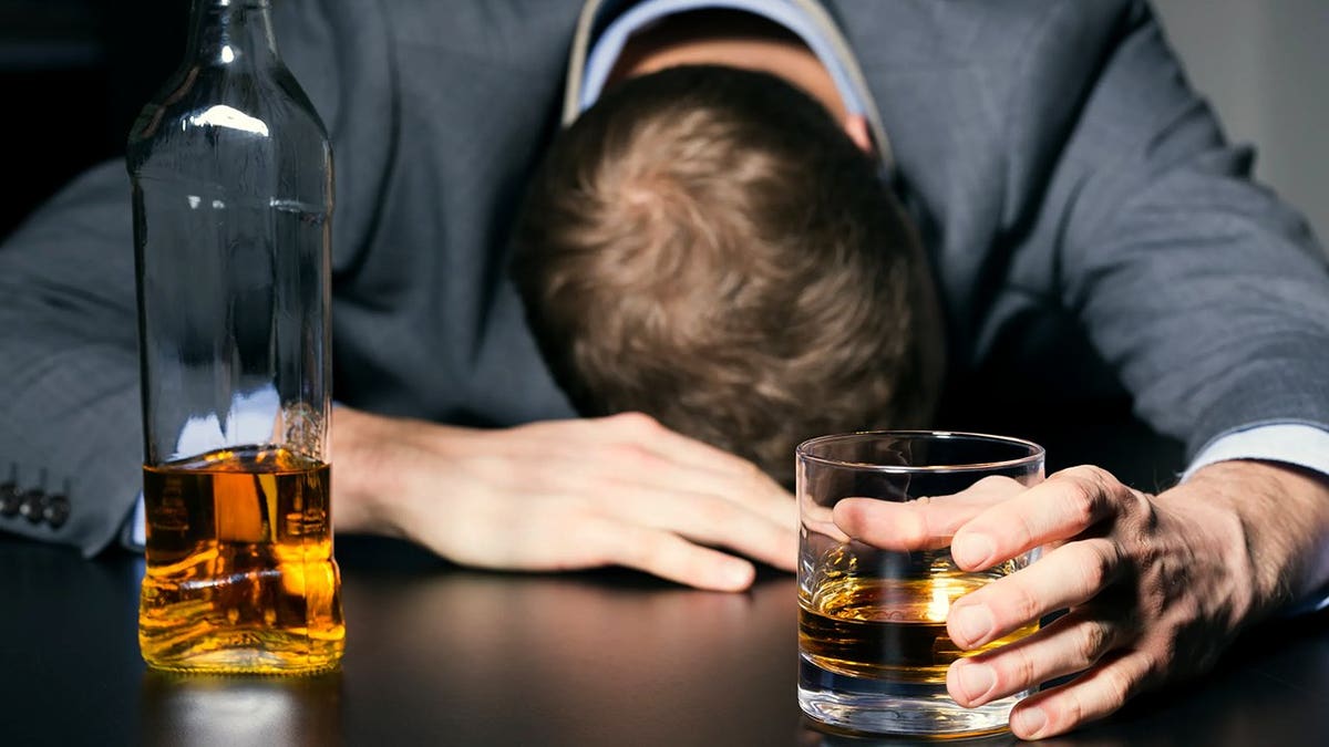 A man seen with head down on bar and hand around glass of liquor with bottle nearby.