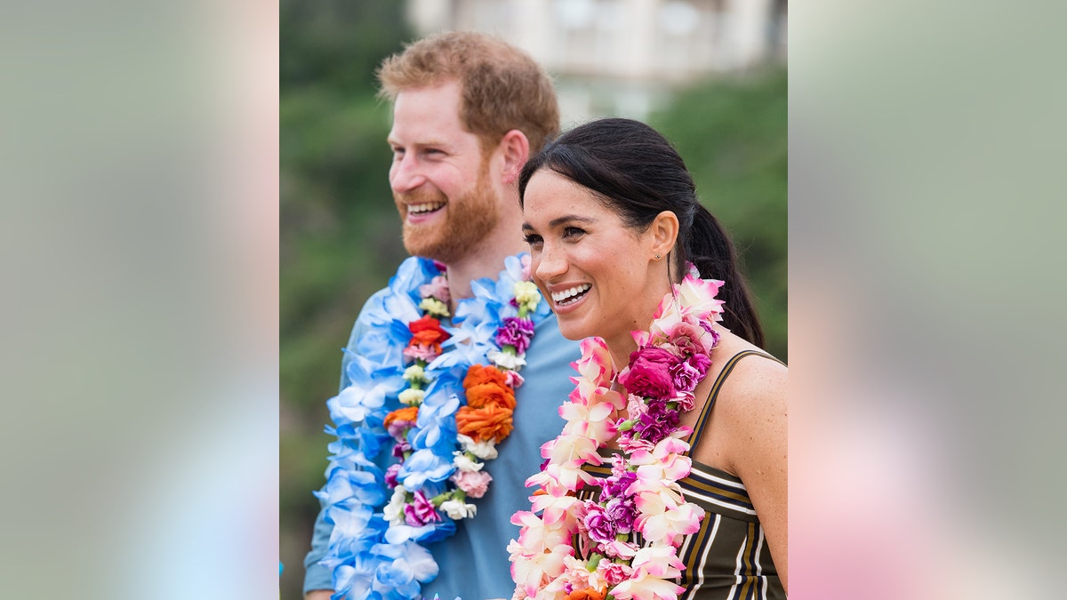 Prince Harry and Meghan Markle smiling with flower wreaths around their necks.