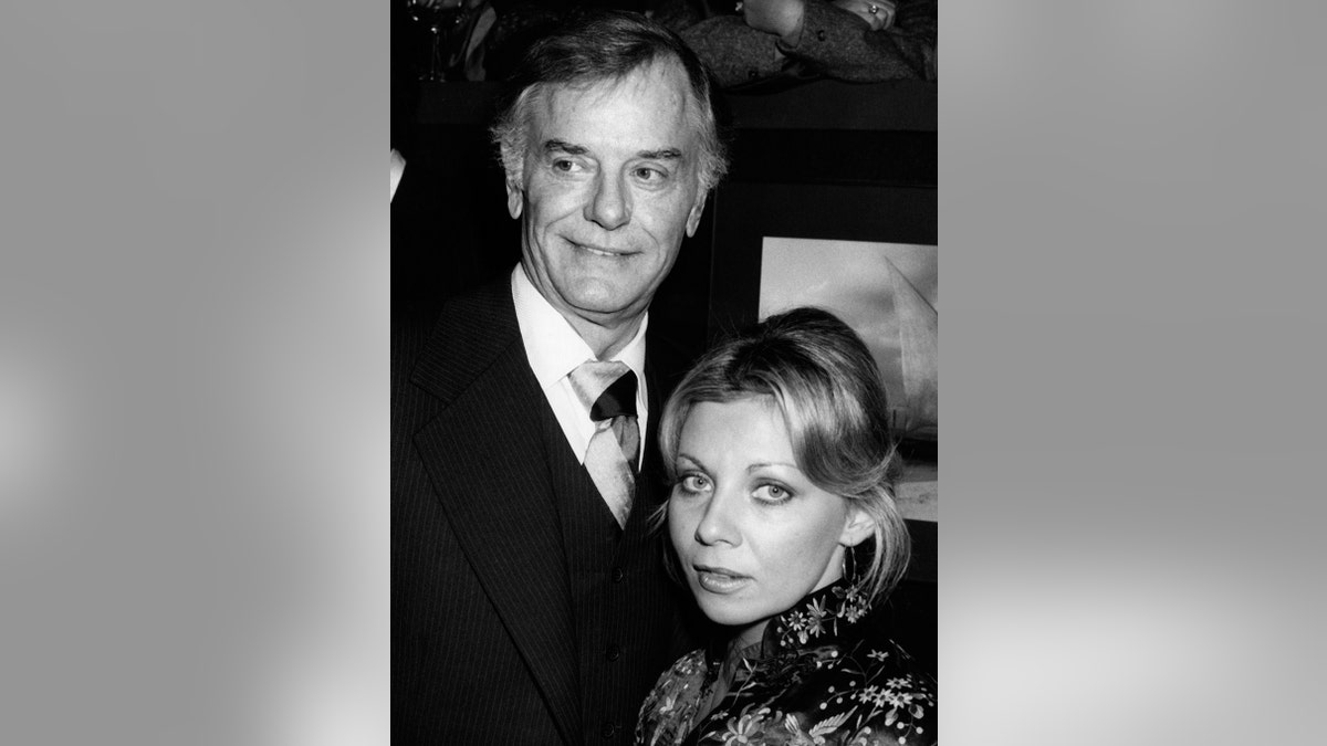 Gig Young smirking in a dark suit next to his wife Kim.