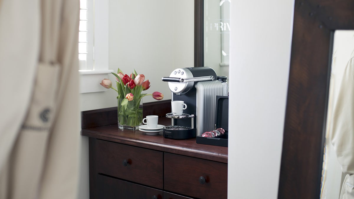 Coffee maker seen on dresser in hotel room with flowers in a vase beside it.