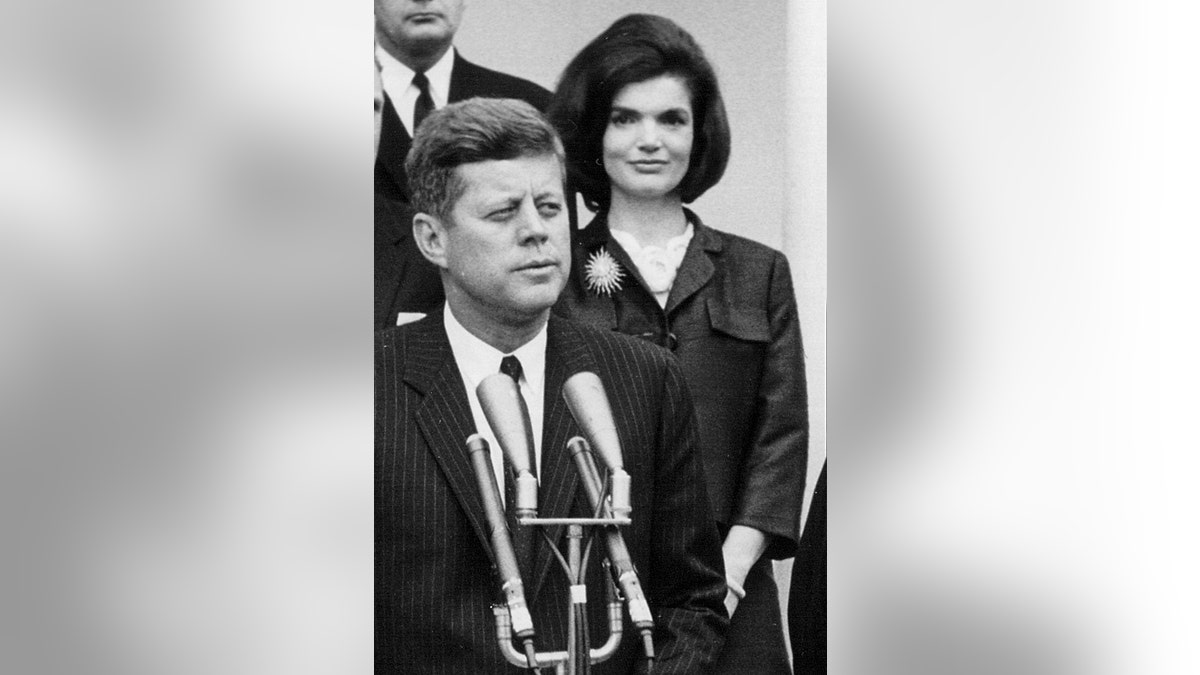 John F. Kennedy speaking at a podium as Jackie Kennedy watches.