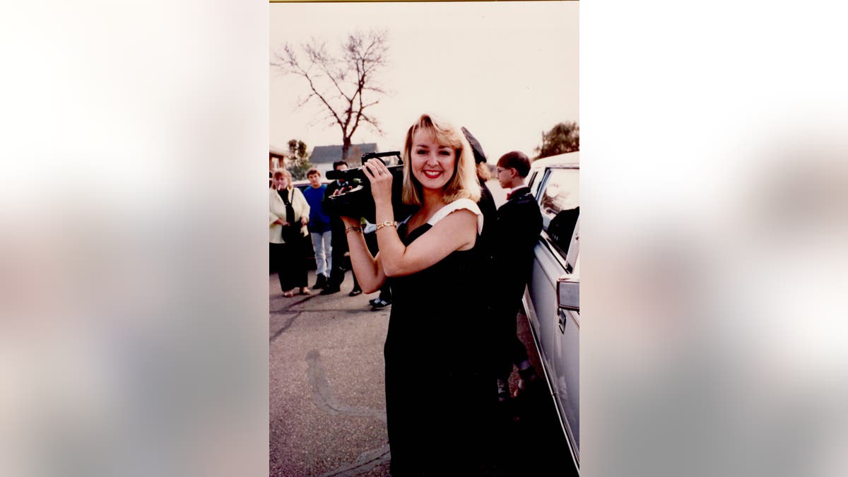 Jodi Huisentruit holds a camera in a black dress and smiling.