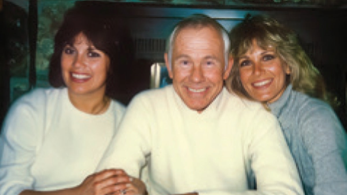 Johnny Carson sonriendo entre dos mujeres sonrientes.