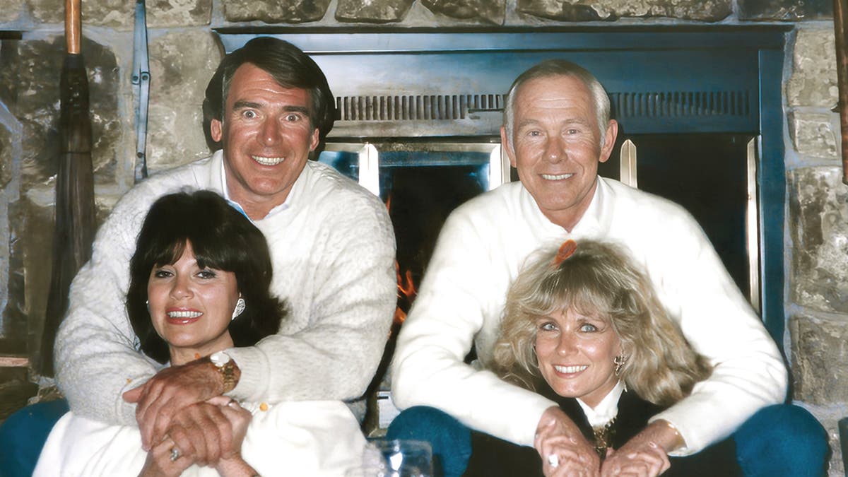 Howard Smith y Johnny Carson con suéteres blancos abrazando a sus esposas.