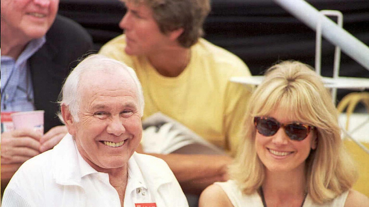 Johnny Carson con una camisa blanca, sentada junto a su esposa con una multitud y sonriendo.