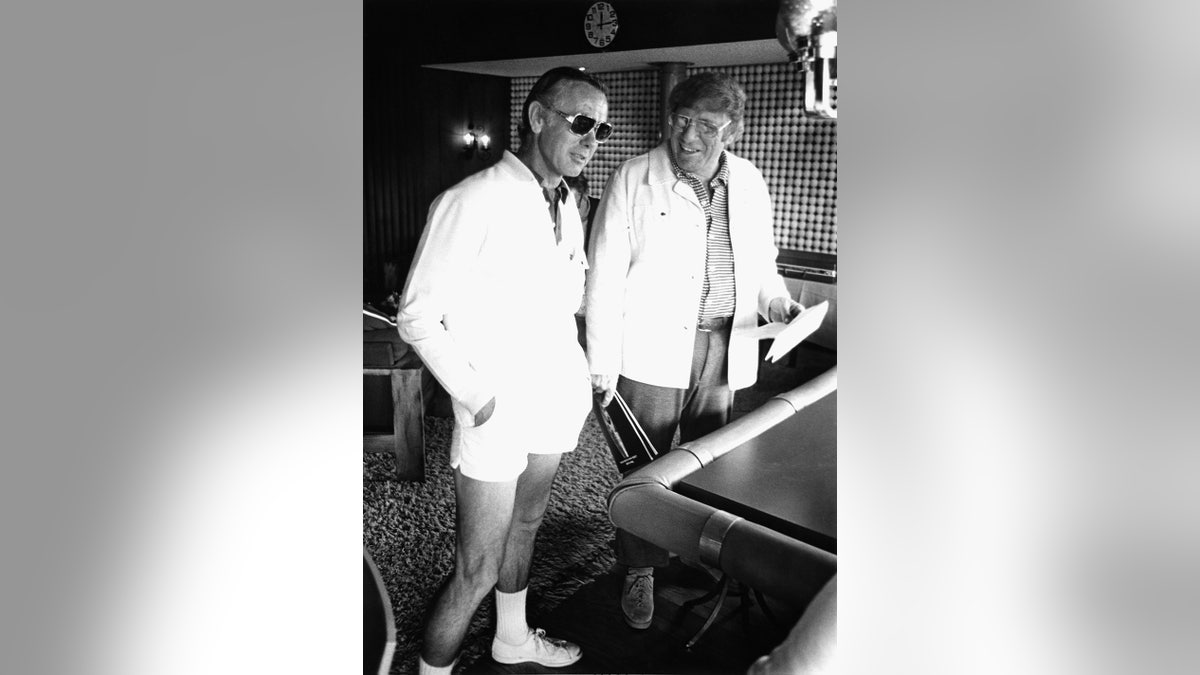 Johnny Carson con un uniforme de tenis con gafas de sol charlando con un amigo.