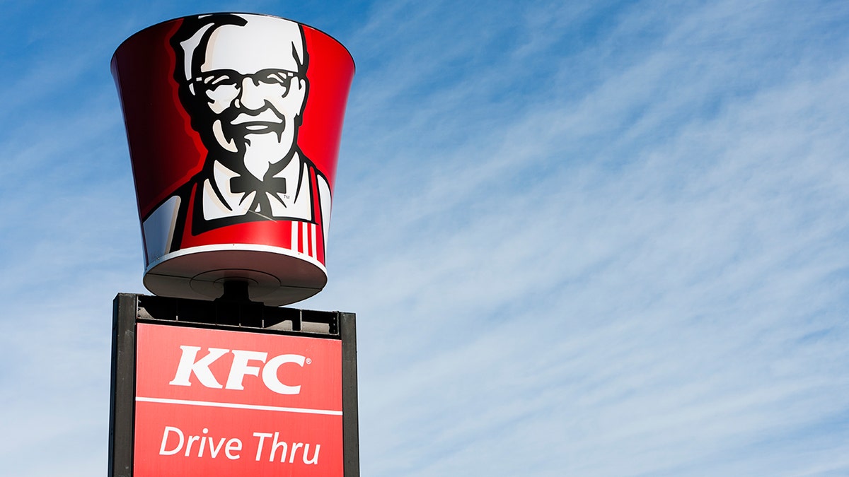 An image of Colonel Sanders is shown on a bucket-shaped sign above the KFC logo behind a blue sky.