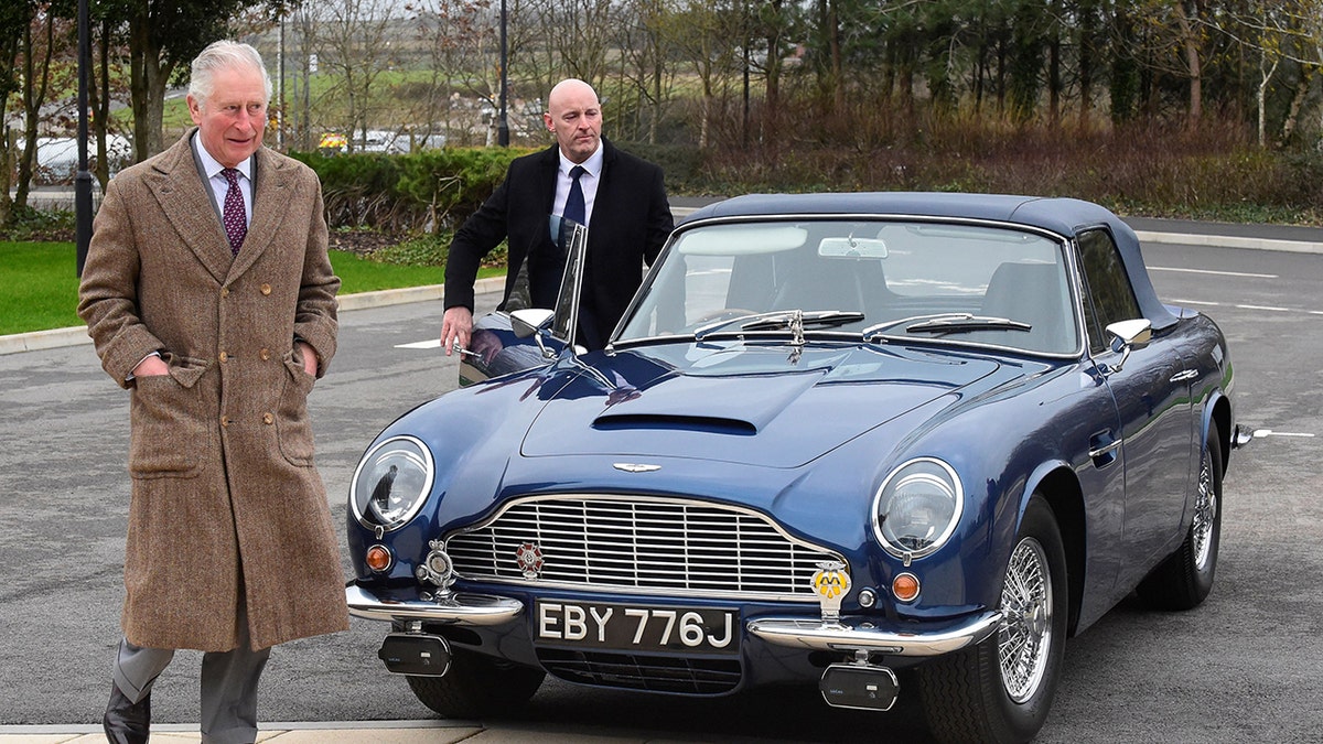 King Charles walking ahead of his car wearing a brown coat and a suit.