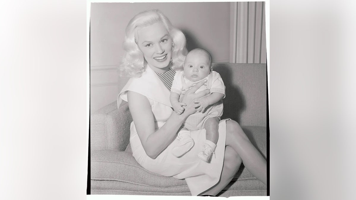 Mamie Van Doren in a white dress smiling holding a baby.