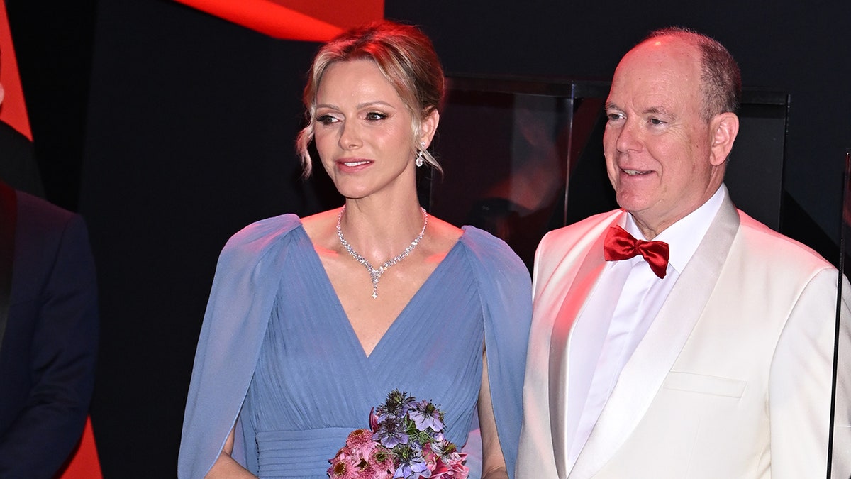 Princess Charlene wearing a flowy bluish dress and holding a bouquet of flowers opinionated adjacent to Prince Albert wearing a achromatic suit pinch a reddish bowtie.