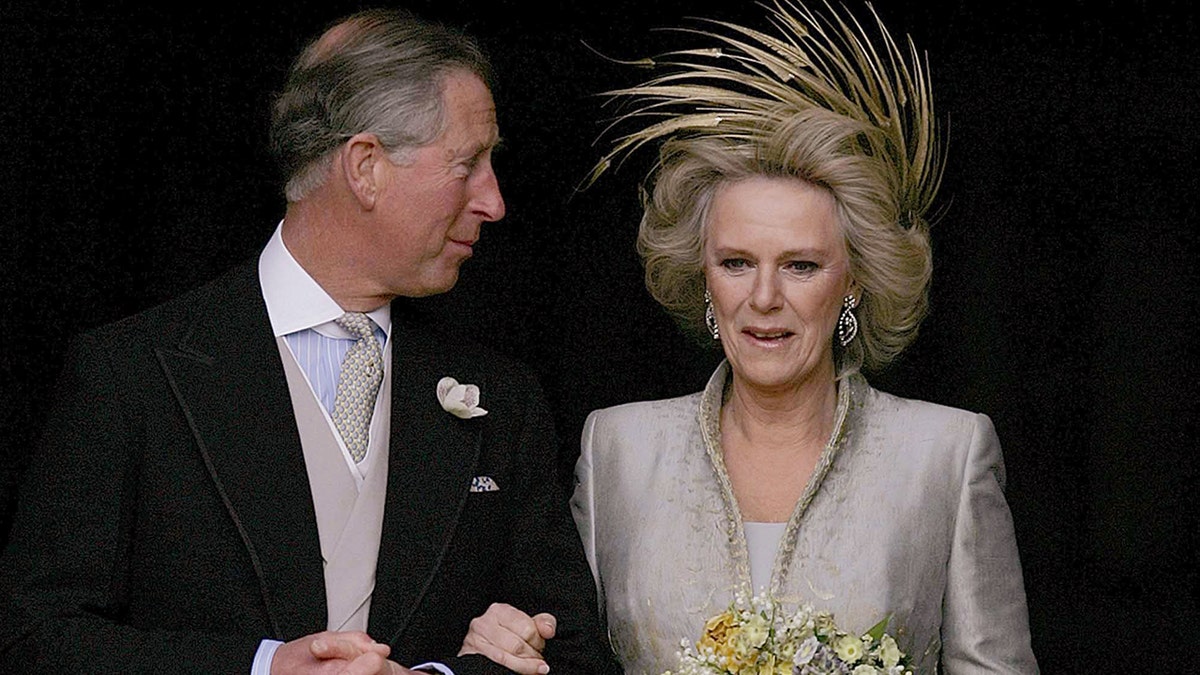 Čārlzs melnā smokā un Kamilla smilškrāsas kleitā un zelta fascinators, kas tur ziedu pušķi.