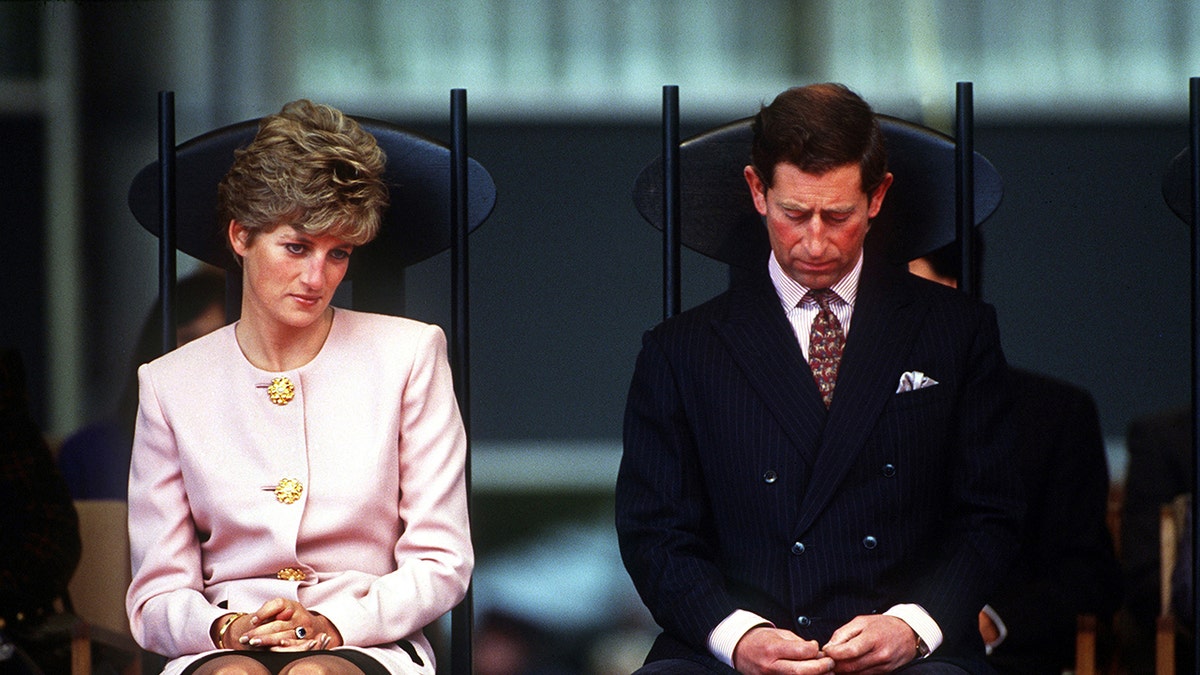 King Charles looks down at the black suit, as Princess Diana in a pink suit looks at her.