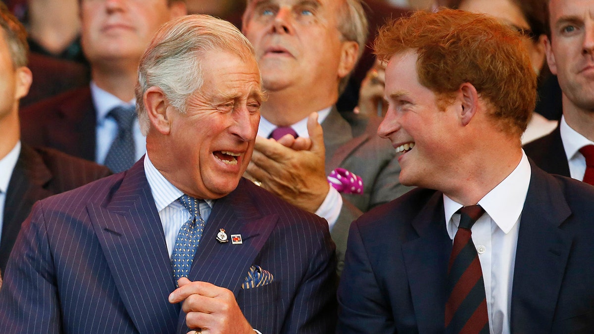 King Charles laughing with his son Prince Harry while sitting at an audience.