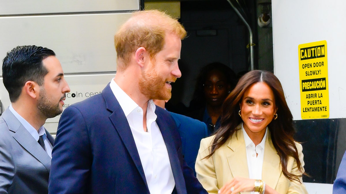 Prince Harry in a blue blazer and white shirt walking next to Meghan Markle in a beige jacket and white shirt.