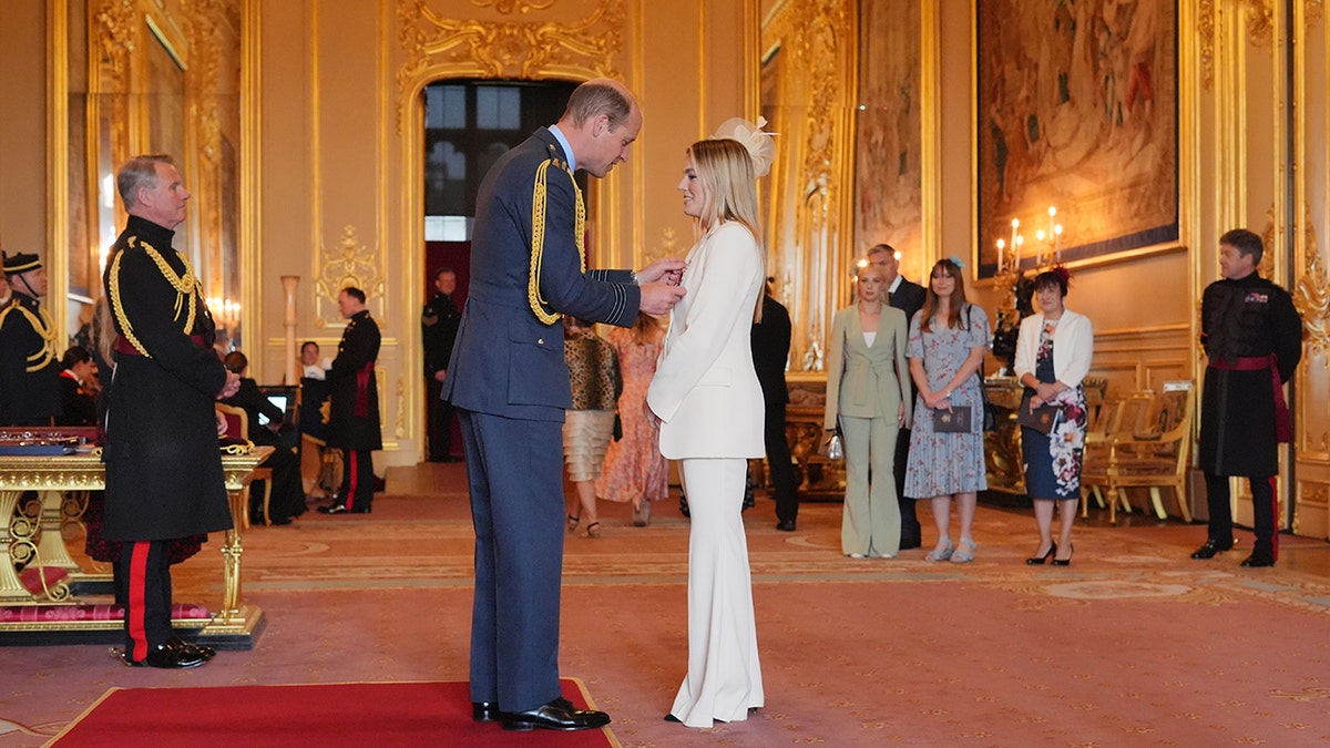 Prince William in a blue suit pinning a medal to a woman who is wearing a white suit.