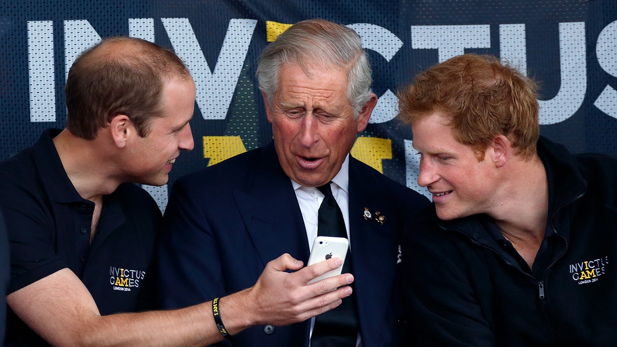 Prince William showing King Charles his phone as Prince Harry looks on.