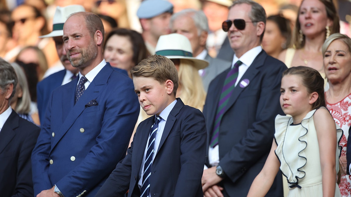 Prince William in a blue suit stands next to his two children.