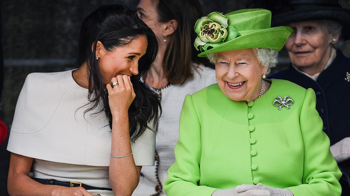 Meghan Markle wearing a beige dress laughing with Queen Elizabeth wearing a bright green dress.
