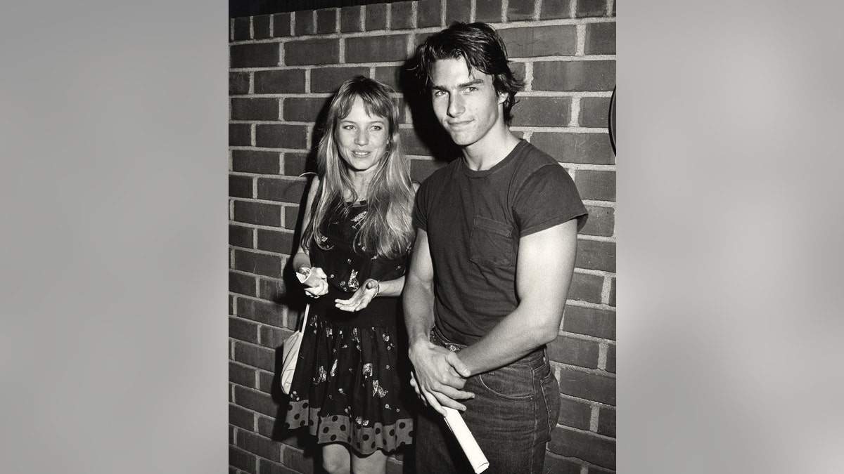 A black and white photo of a young Rebecca De Mornay and Tom Cruise standing against a brick wall.