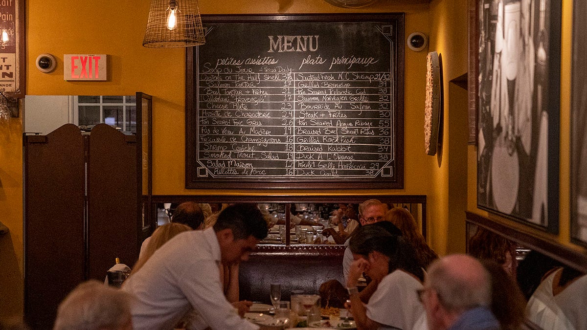 Large chalkboard menu seen at the back of a restaurant filled with customers, servers working.