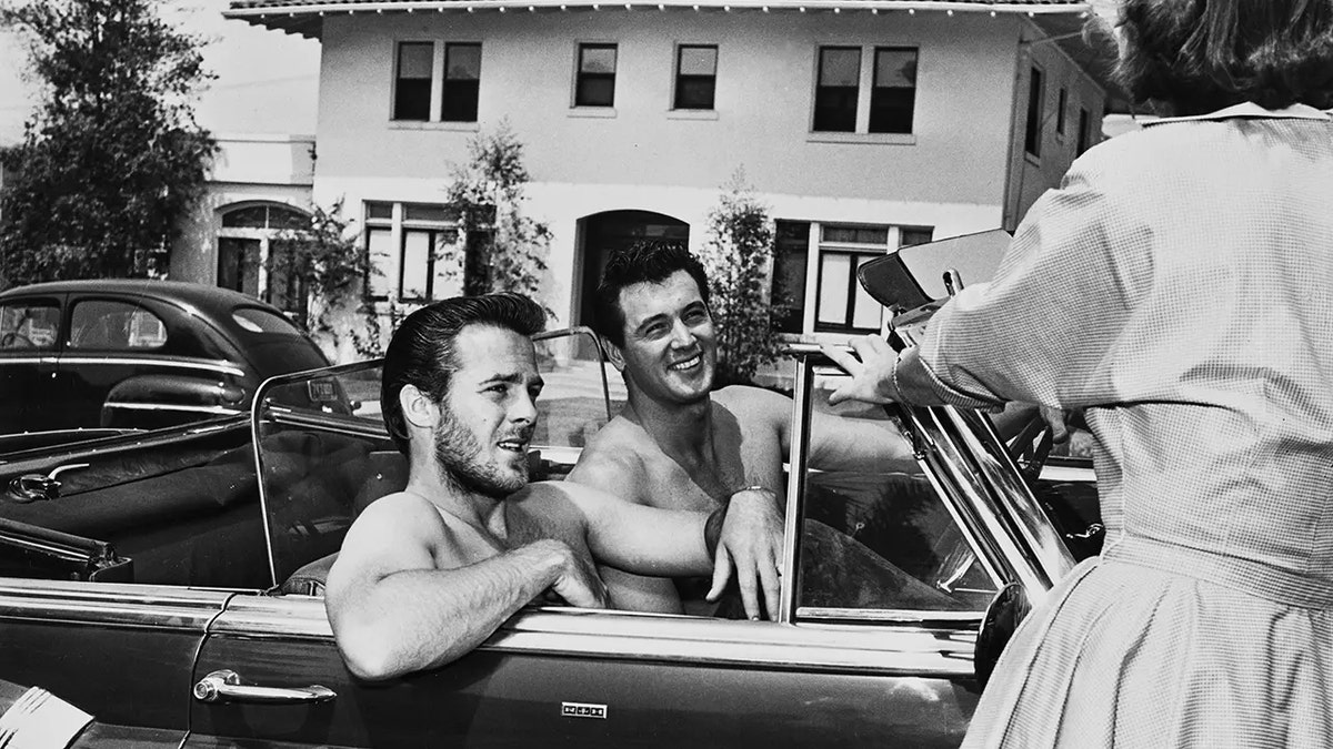 Rock Hudson smiling in a car sitting next to a man while looking at a woman.