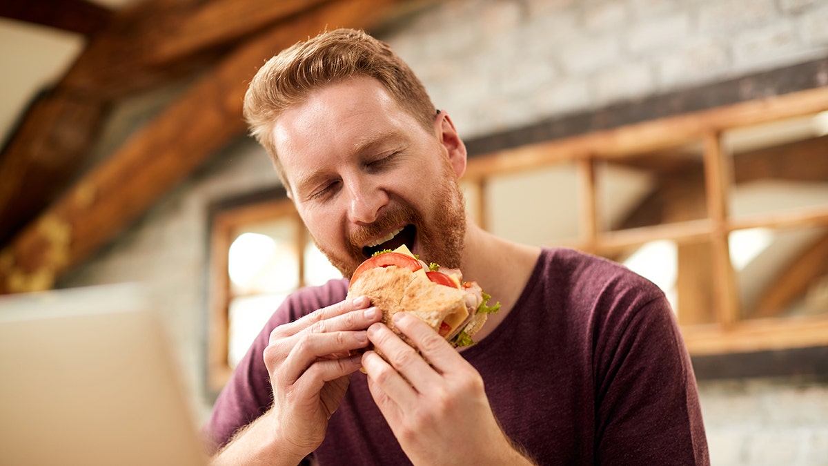 Man with eyes closed eating sandwich for breakfast at home.