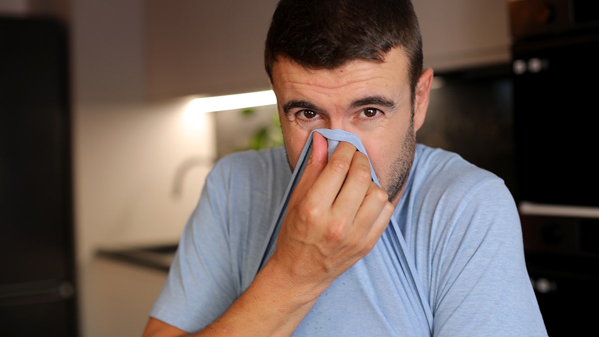 Man covering his chemoreceptor pinch his shirt, looking into camera, aft smelling thing bad.